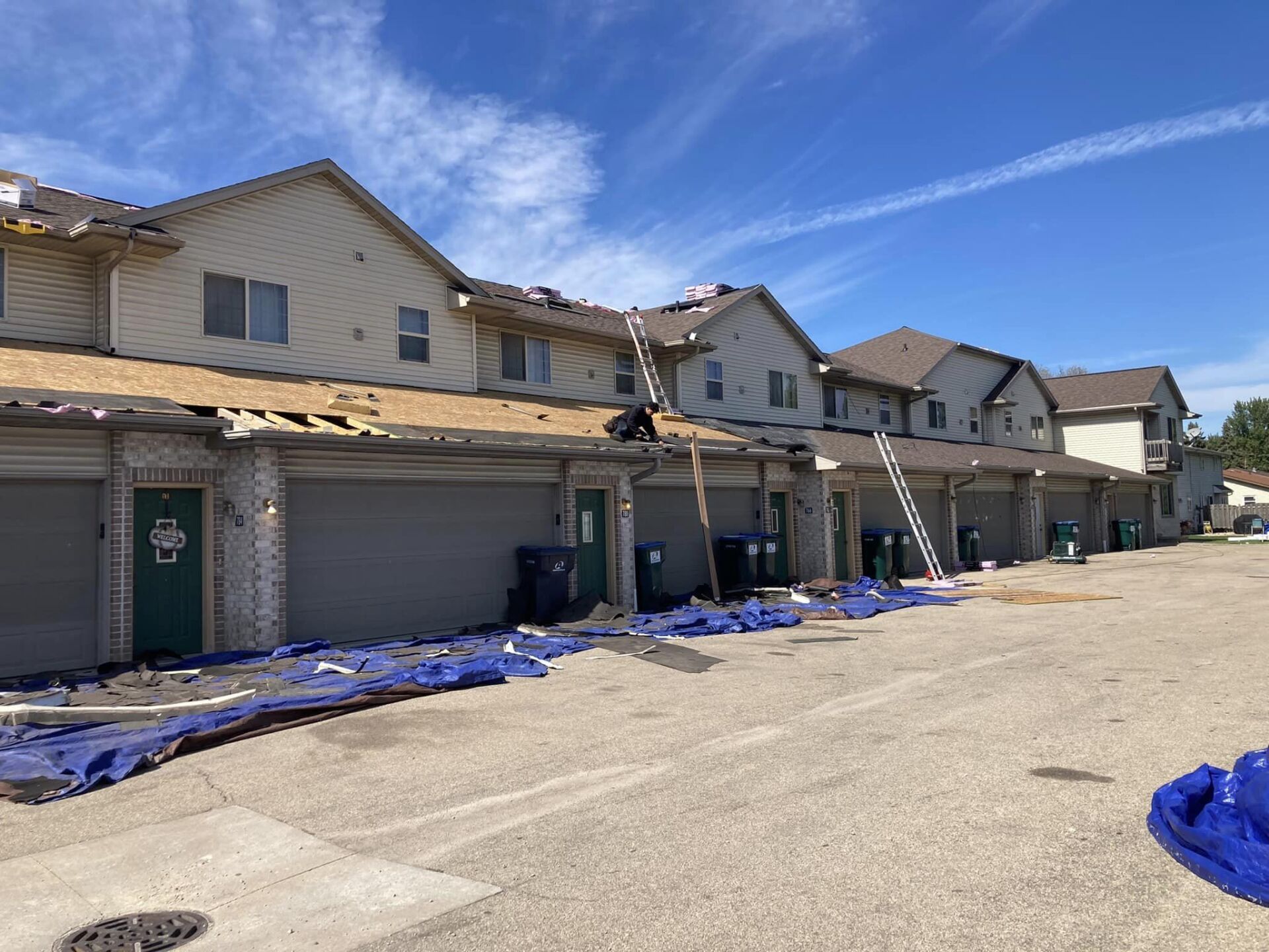 Workers perform roof repairs on a row of townhouses with beige siding, featuring a ladder and blue tarps on the ground.