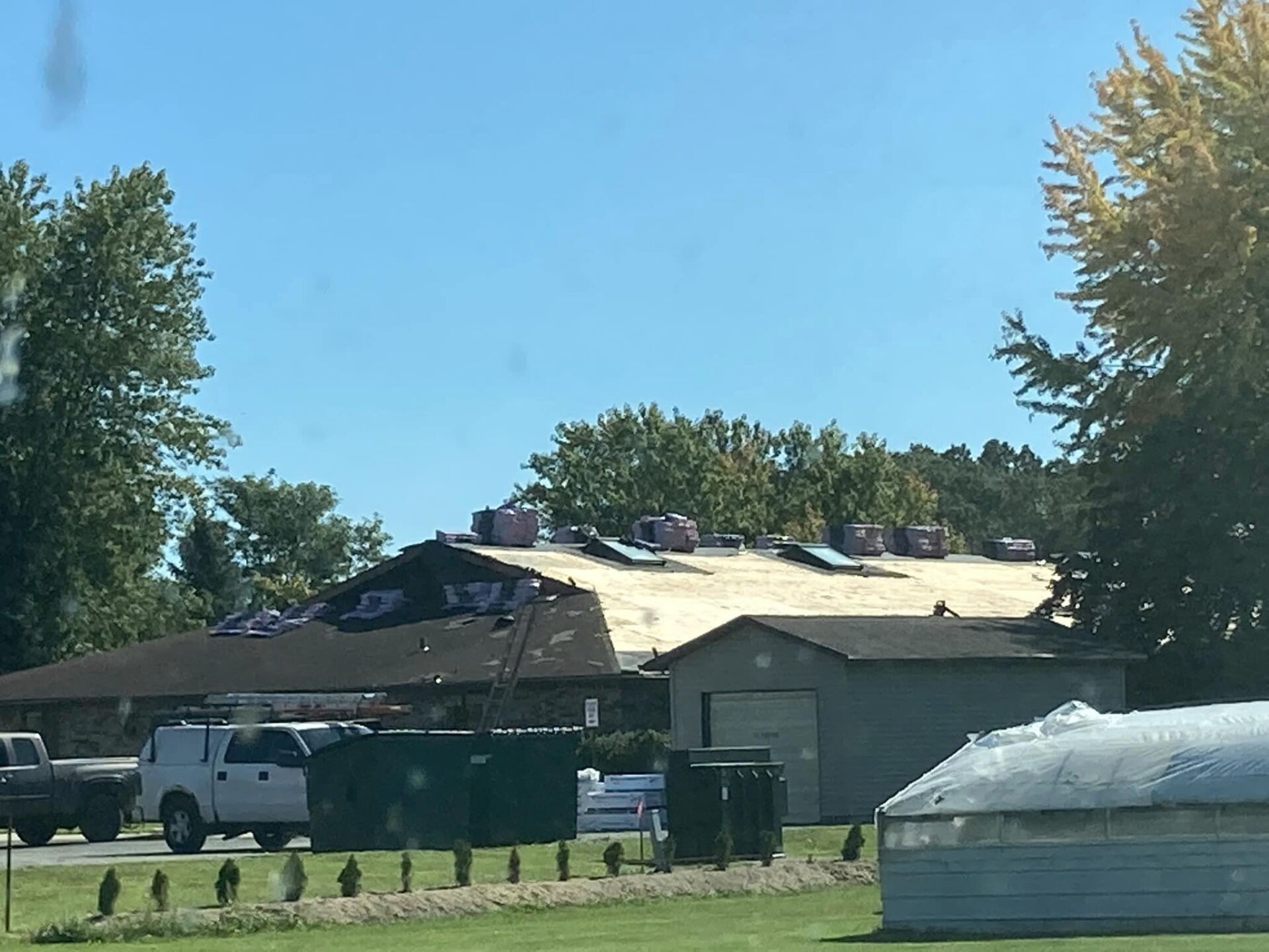A building under construction with a partially shingled roof, surrounded by a parking lot, a dumpster, and green trees.