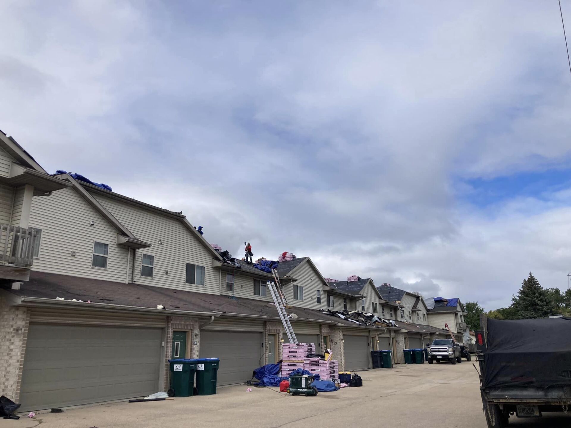 Workers performing roof repairs on a row of townhomes with garages, a ladder, and construction materials on the ground.