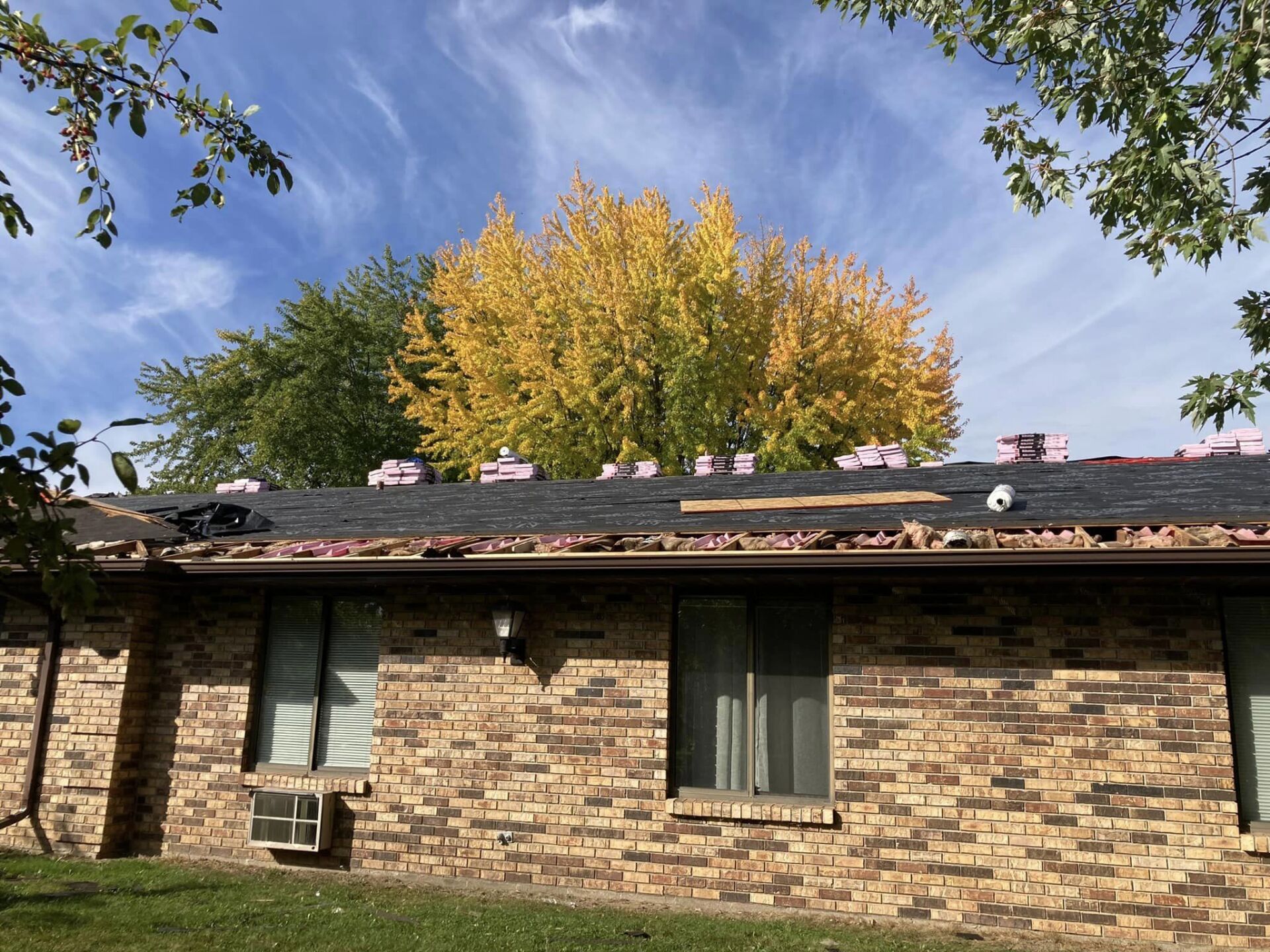 A brick house with a partially removed roof during a roofing project, framed by trees with green and autumn yellow leaves.