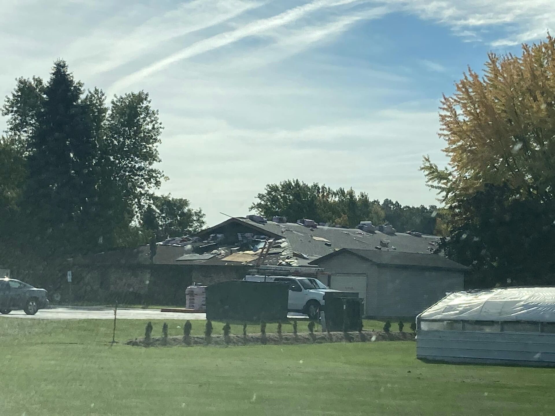 A damaged residential building with significant roof destruction, surrounded by trees, a parked truck, and a grassy yard.