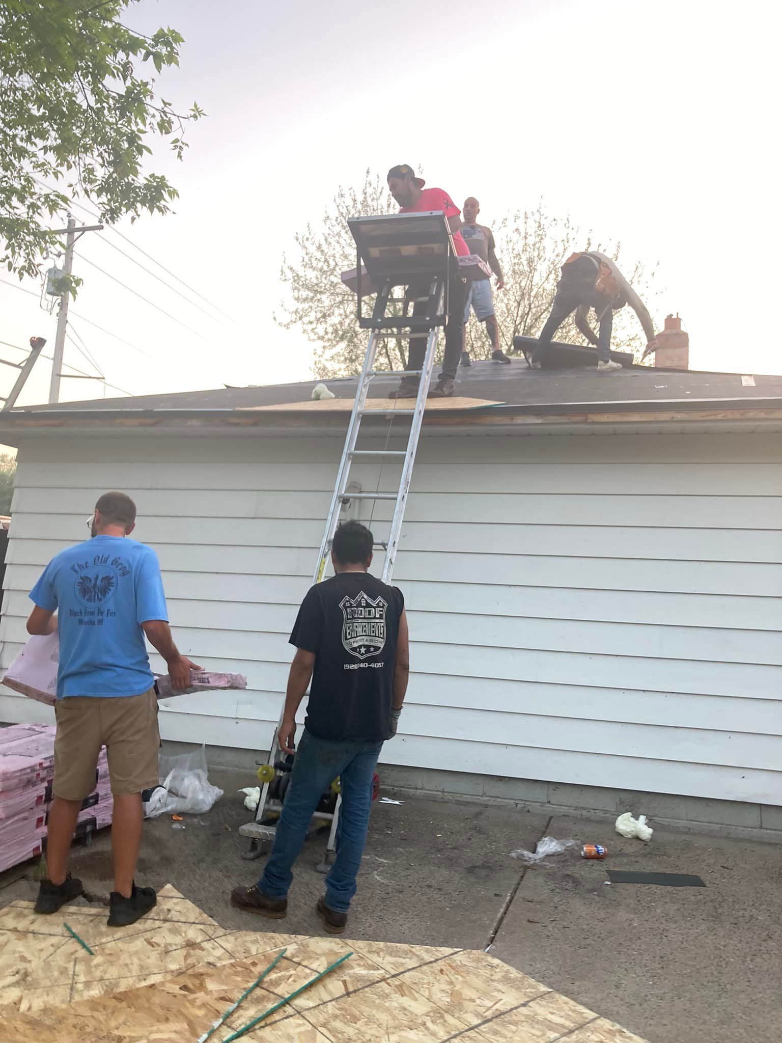 Four workers replace a shingled garage roof using a ladder hoist and OSB panels on a sunny day.