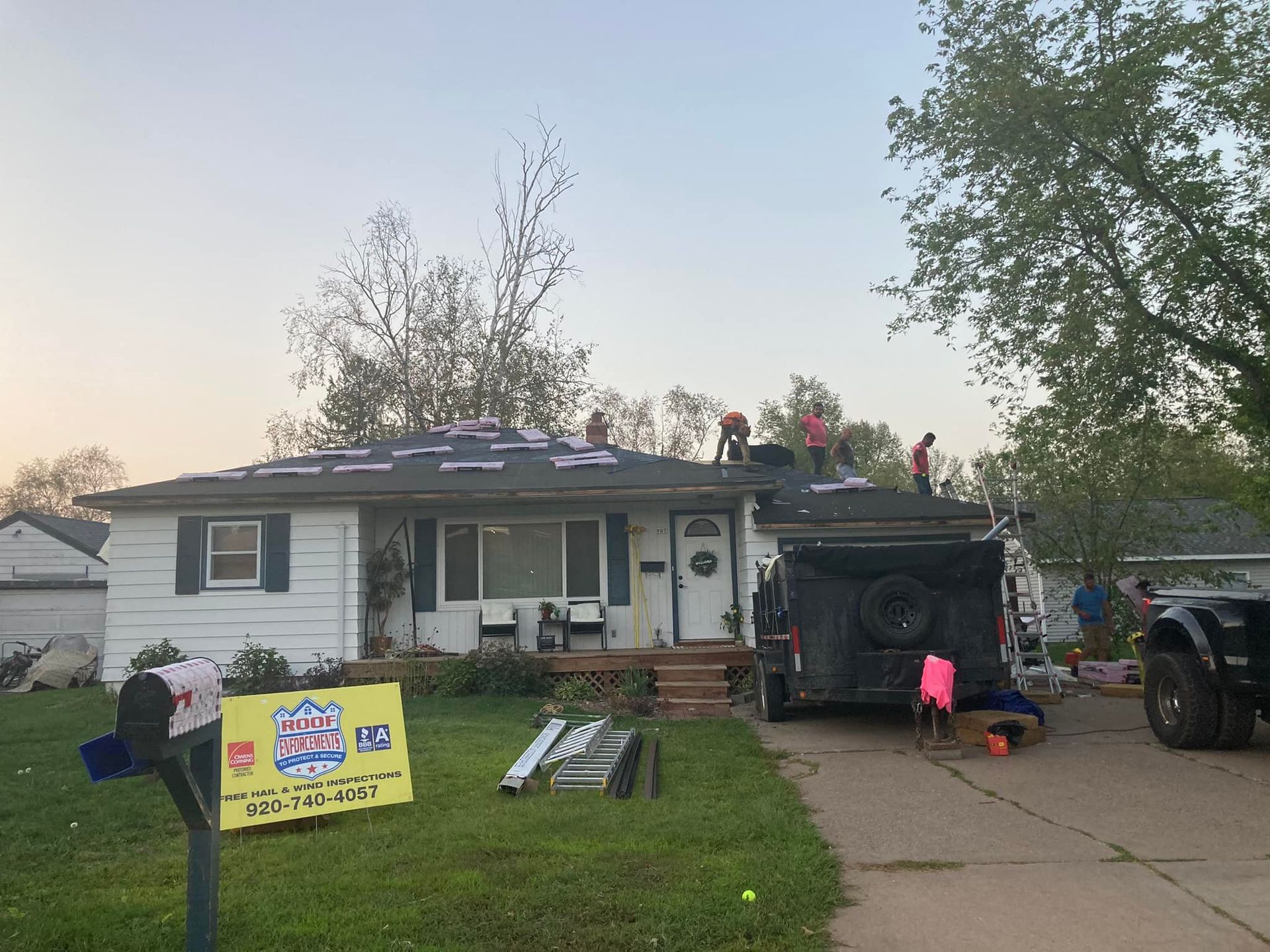 Workers install a new roof on a white, single-story suburban house with a sign in the front yard.