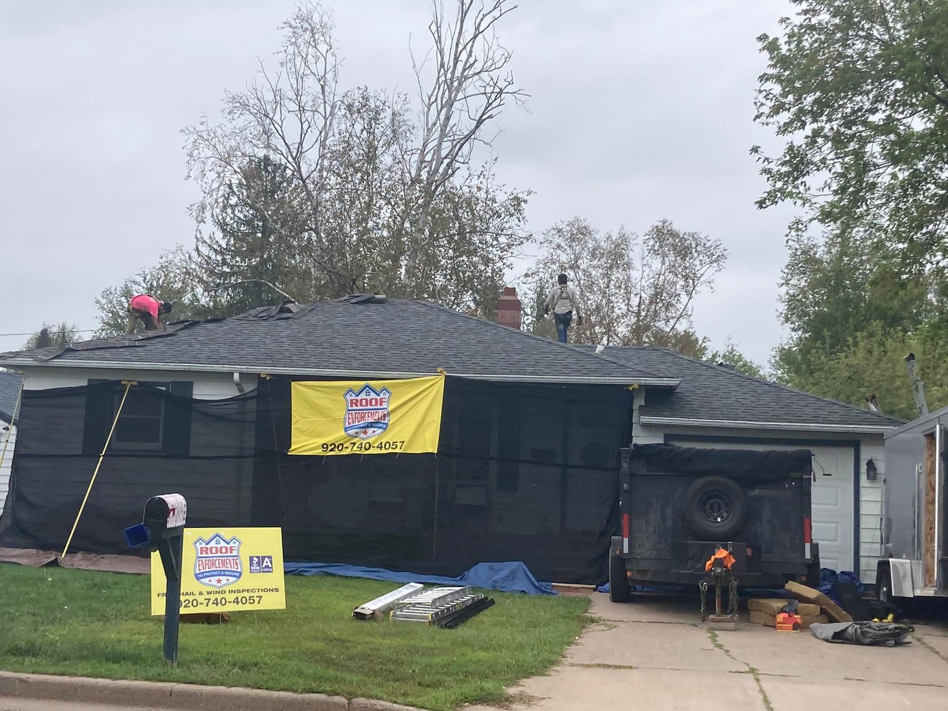 Workers on the roof of a house wrapped in black safety netting, with promotional banners displayed on the lawn and porch.