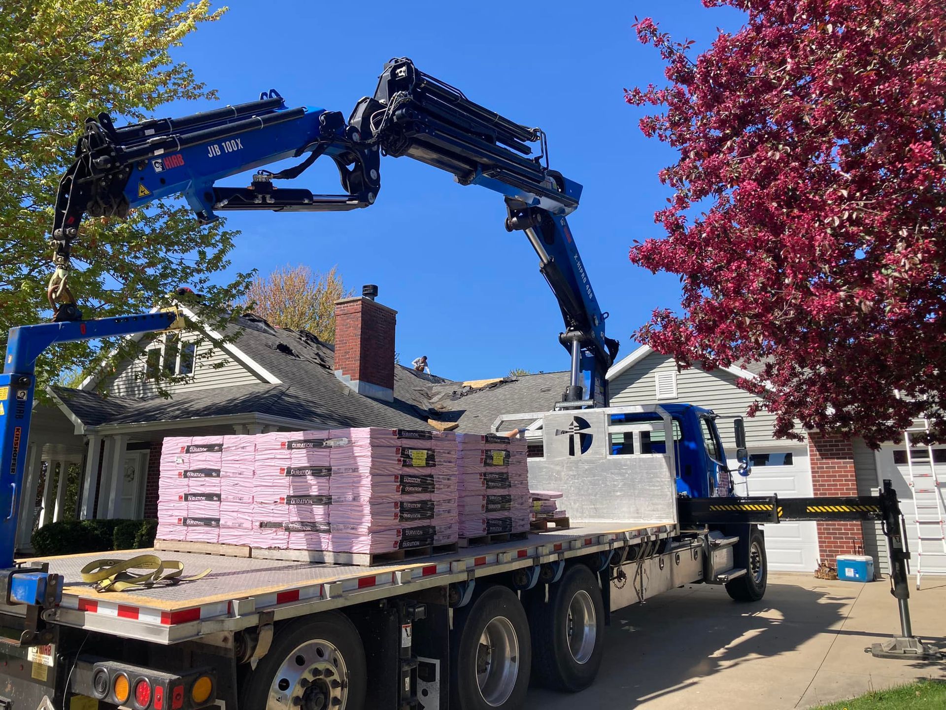 A blue boom truck with a crane arm parked on a residential driveway, loaded with stacks of pink foam insulation panels.
