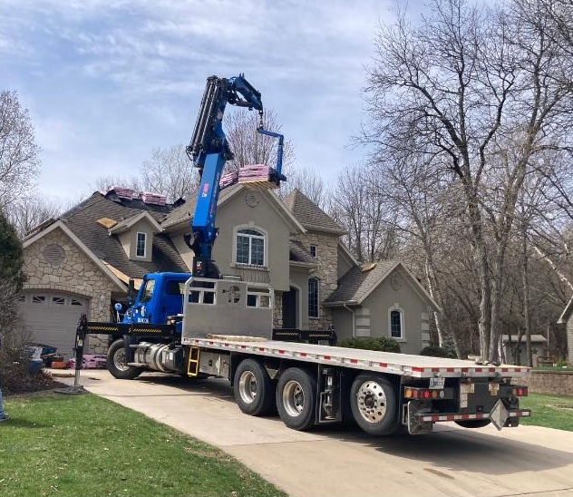 A blue crane truck parked in a suburban driveway lifts roofing materials toward the roof of a large house.
