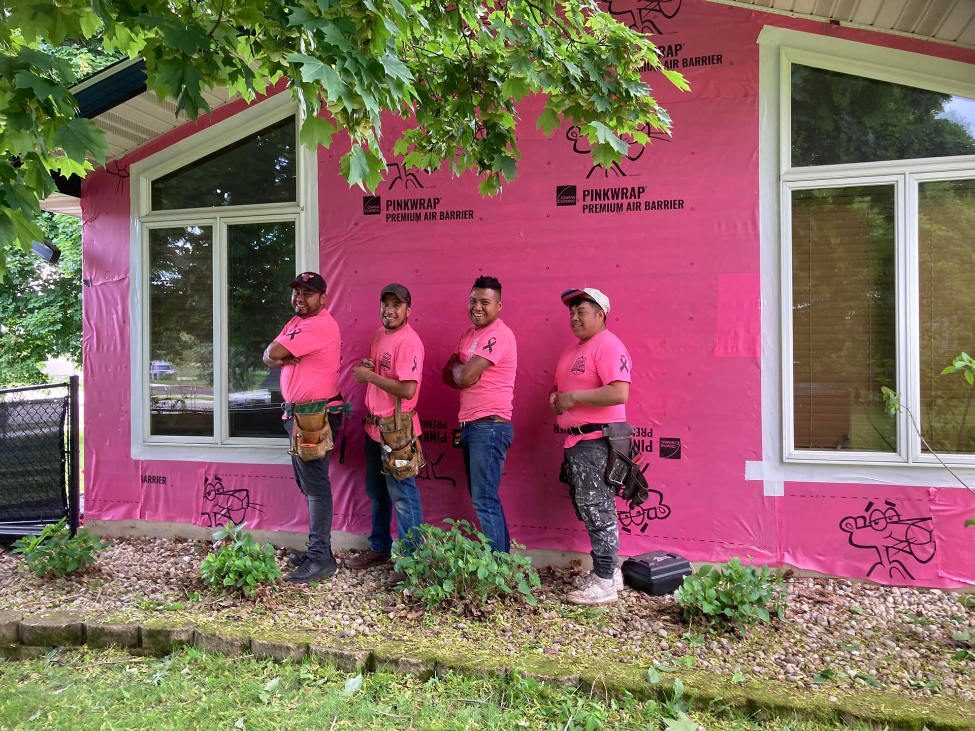 Four workers in matching pink shirts and tool belts stand against a house wall covered in pink protective sheathing.