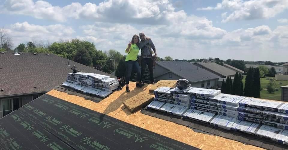 Two people stand on a residential roof under construction, posing next to stacks of shingles on a sunny day.