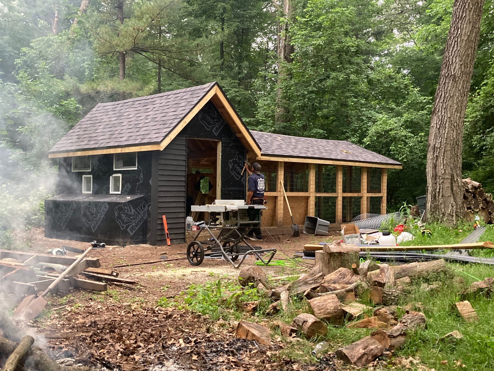 Construction site: a small, dark-sided building with a light wood extension; person working. Surrounded by trees and debris.