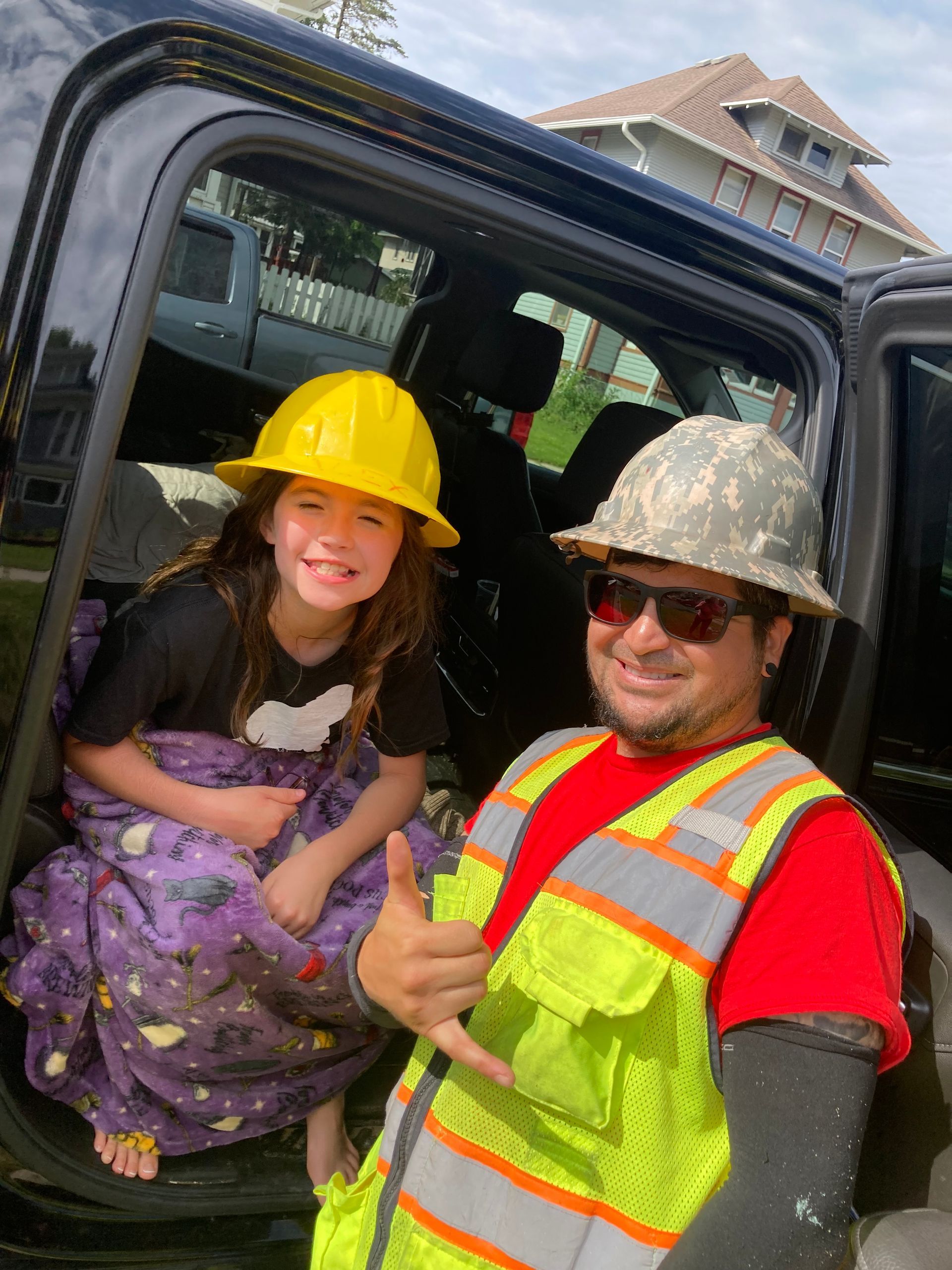 A person in a high-visibility vest and camouflage hard hat gives a shaka sign next to a child wearing a yellow hard hat.