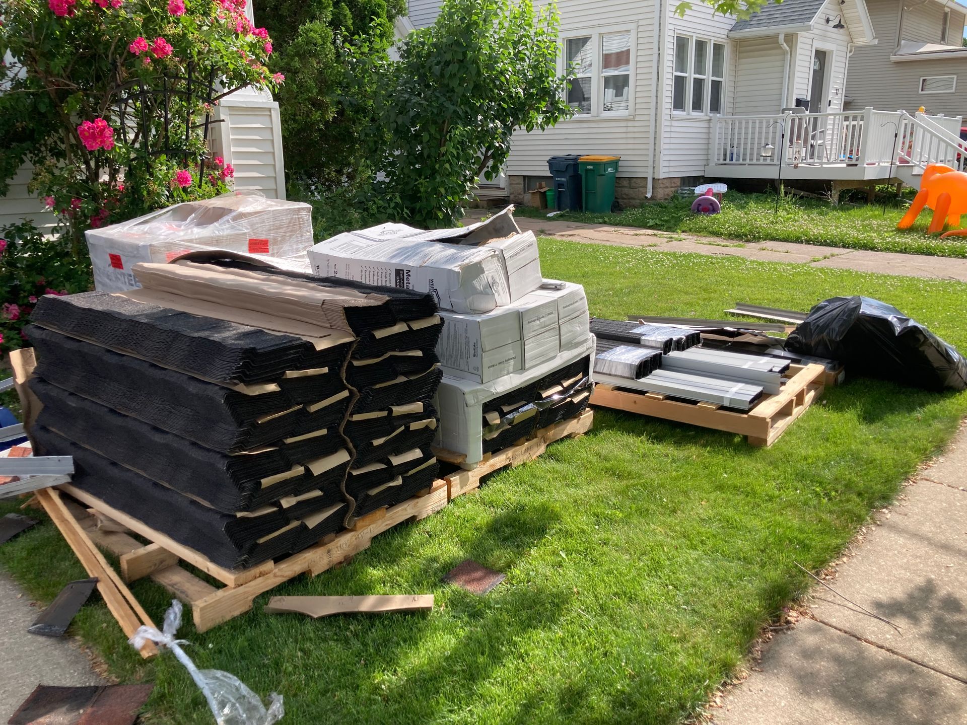 Wooden pallets holding stacks of black roofing shingles and white bundles sit on a grassy lawn in front of a house.