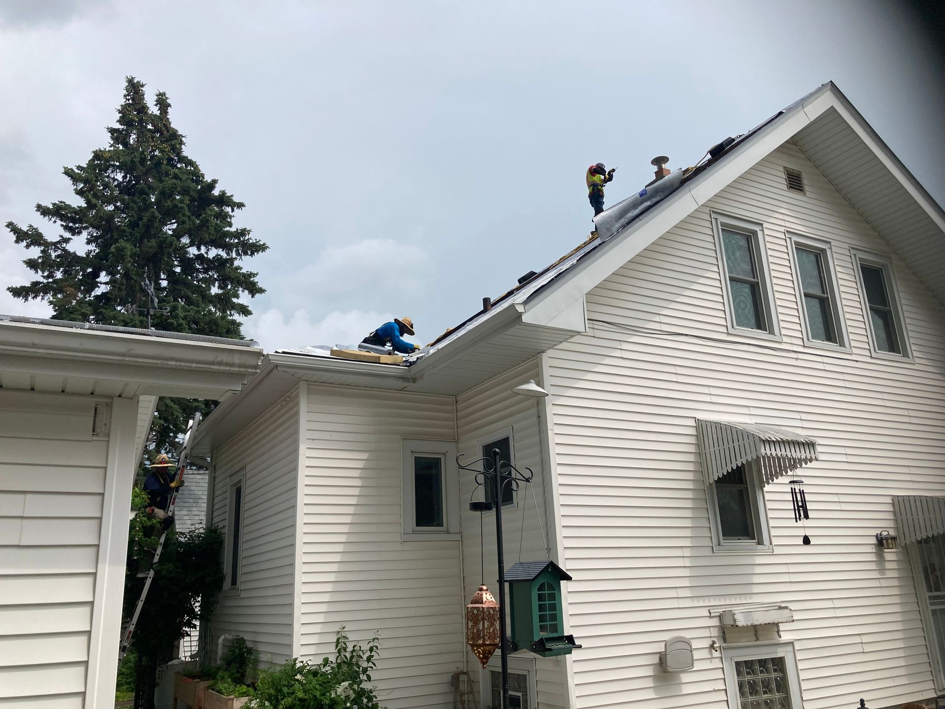 Two people in safety gear work on the roof of a white, two-story house under a cloudy sky.