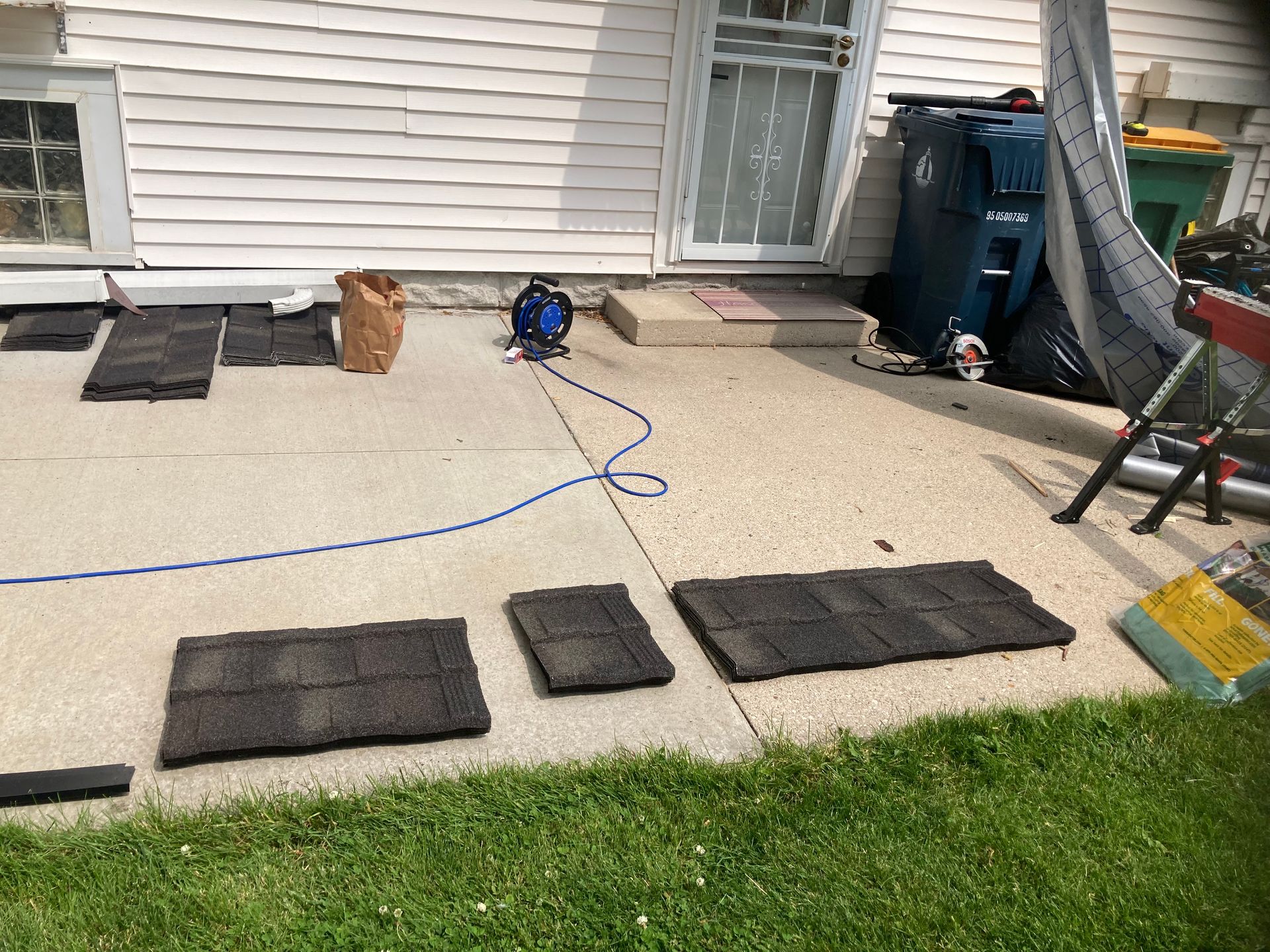 Black roof shingles are laid out in rows on a concrete patio next to a house with a blue extension cord and trash bins.