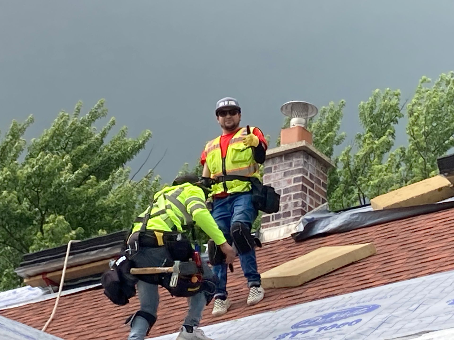 Two construction workers in high-visibility vests and safety gear work on a sloped residential roof near a brick chimney.
