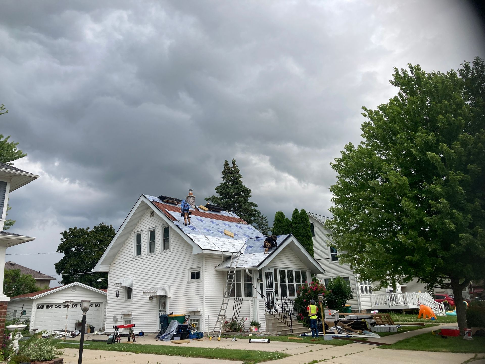 Workers repair the roof of a white house under a cloudy sky, with materials and equipment scattered on the lawn.