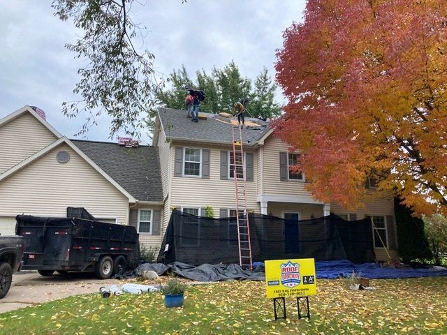 Workers repair the shingled roof of a light-colored, two-story house with a dumpster and yard sign nearby.
