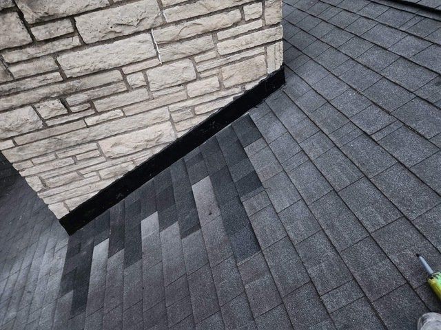 A stone chimney meets a shingled roof with a black, step-flashed seal along the roof-to-chimney connection.