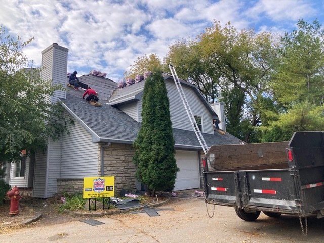 Two workers in red shirts install gray shingles on a multi-story home roof, with a trailer parked in the driveway below.