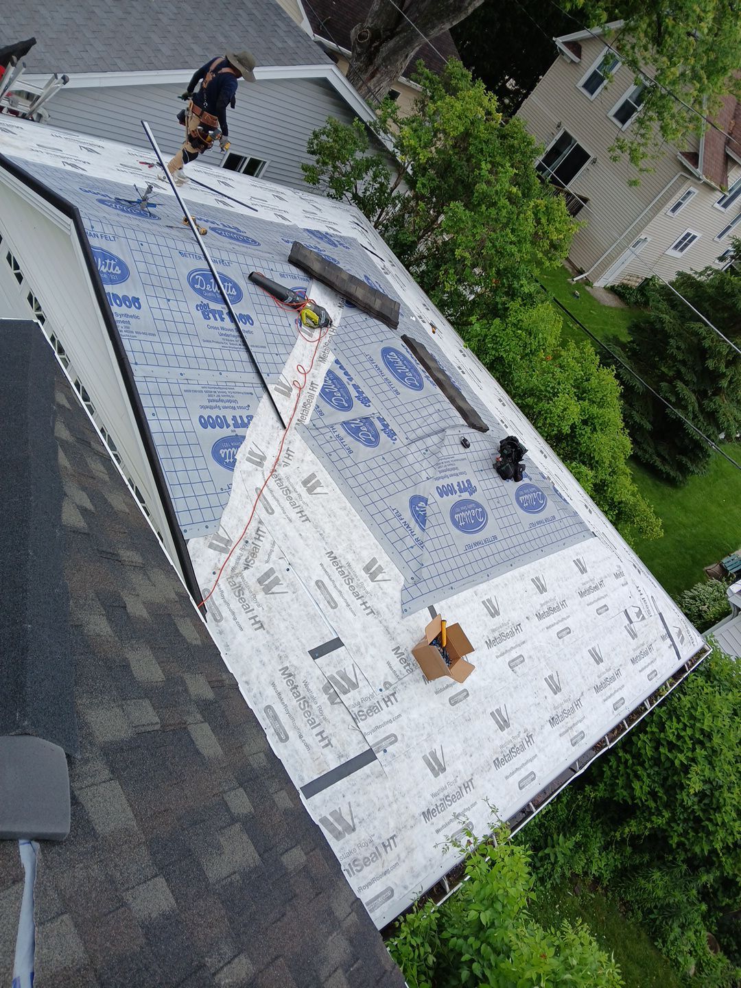 A worker stands on a residential roof partially covered in white underlayment, with a box of tools nearby.