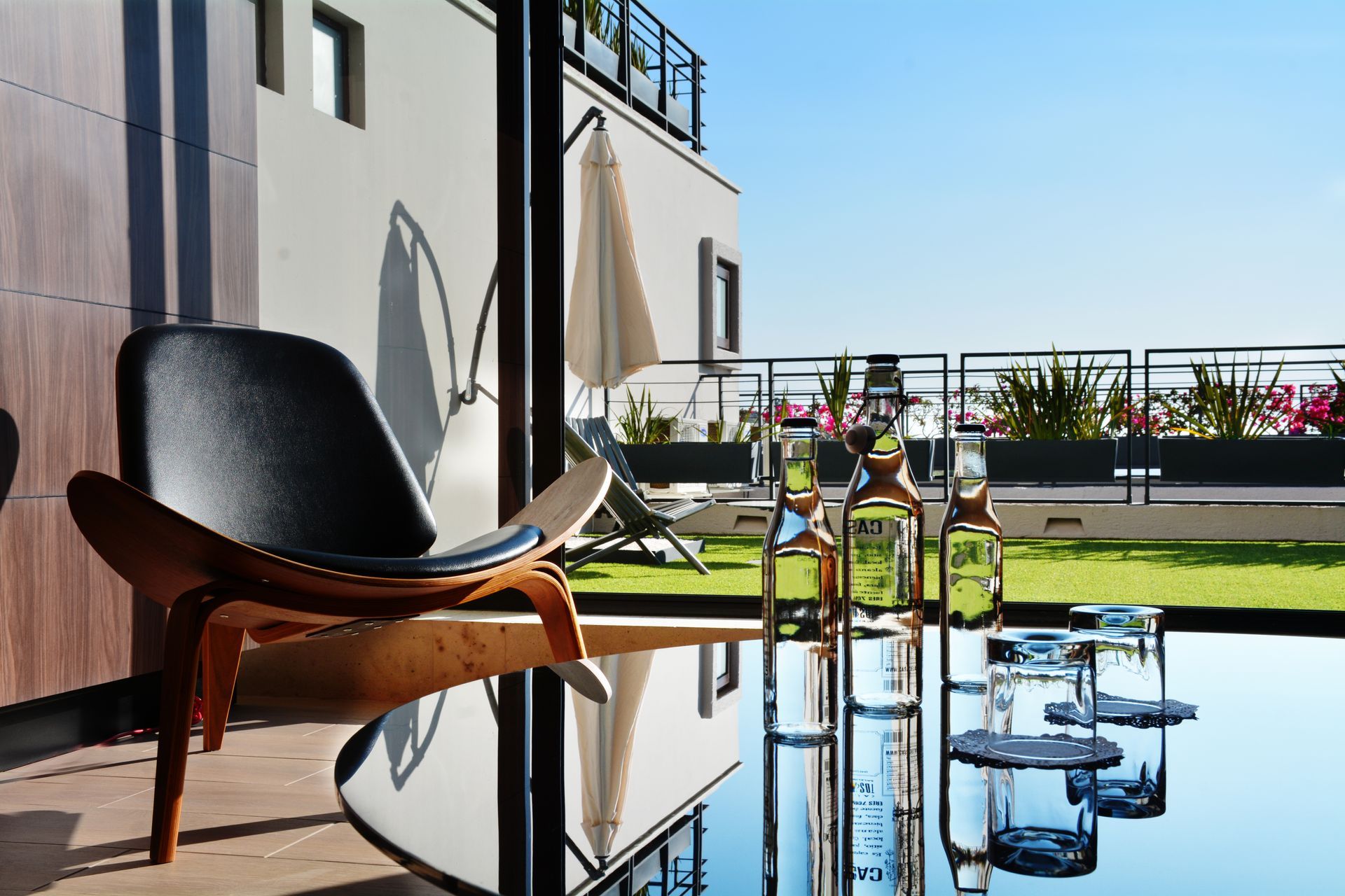 Patio al aire libre con sillas, mesa, bebidas y una vista soleada de un jardín en la azotea y barandillas.