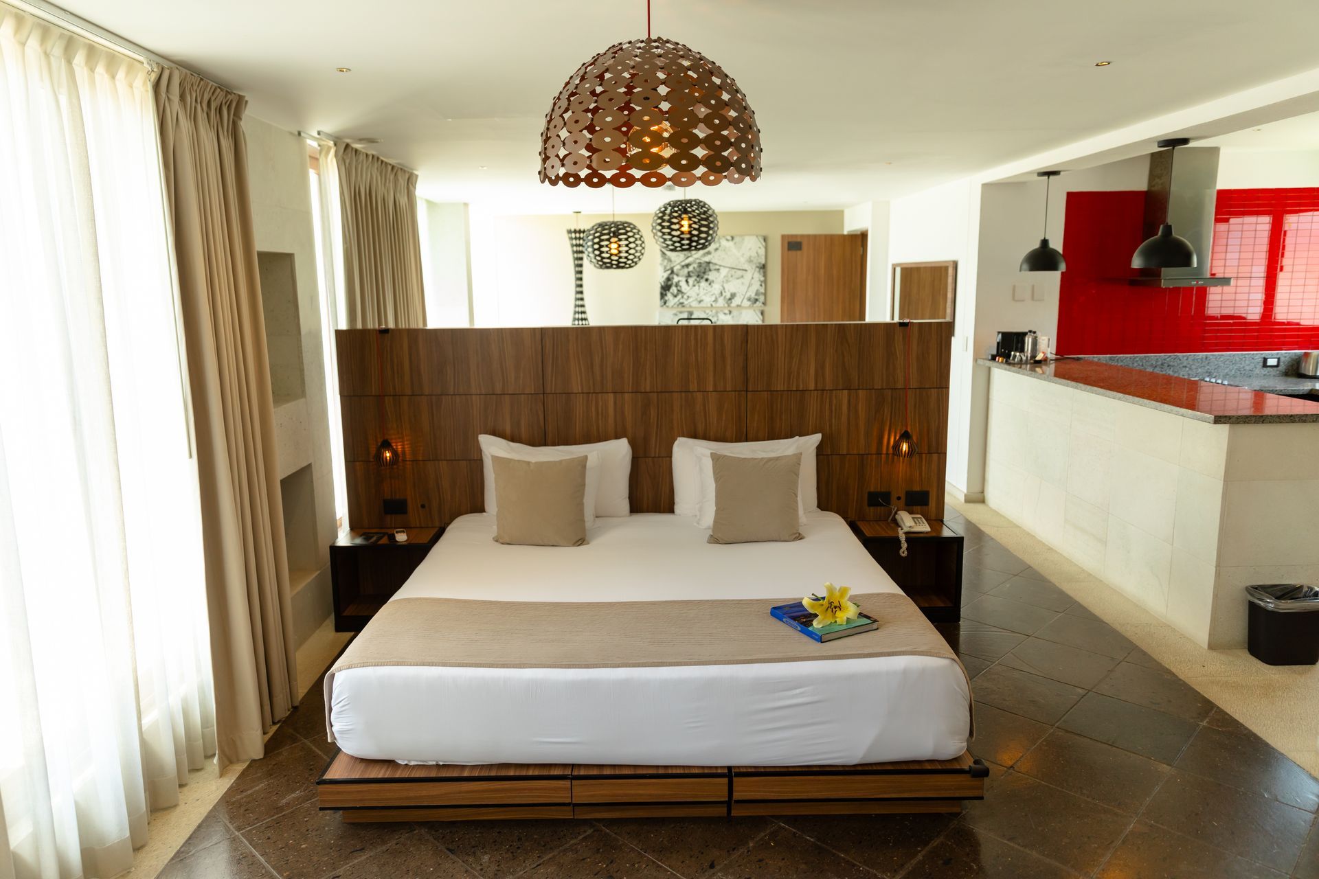 Dormitorio con cama king size, cabecero decorativo y cocina americana. Alfombra oscura sobre suelo de baldosas.