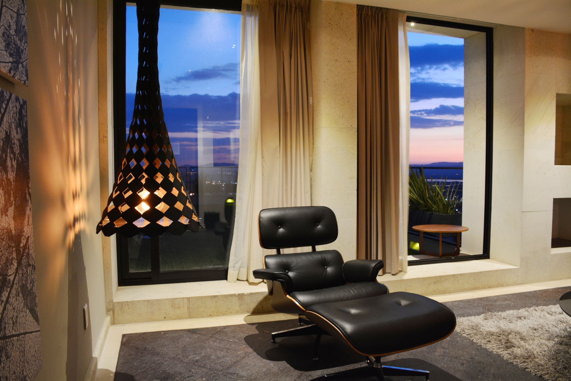 Sillón negro y otomana junto a un gran ventanal, con vistas al atardecer. Lámpara colgante.