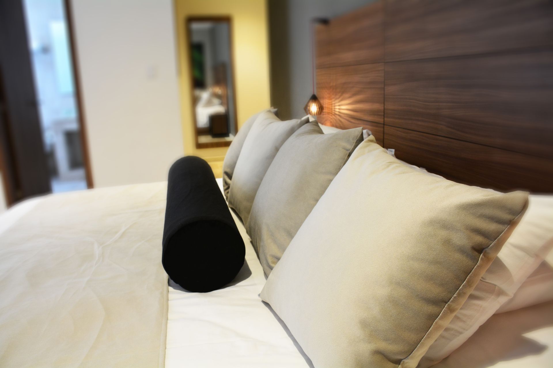 Cama con sábanas blancas, almohadas color canela y un almohadón negro. Al fondo, un cabecero y un espejo.