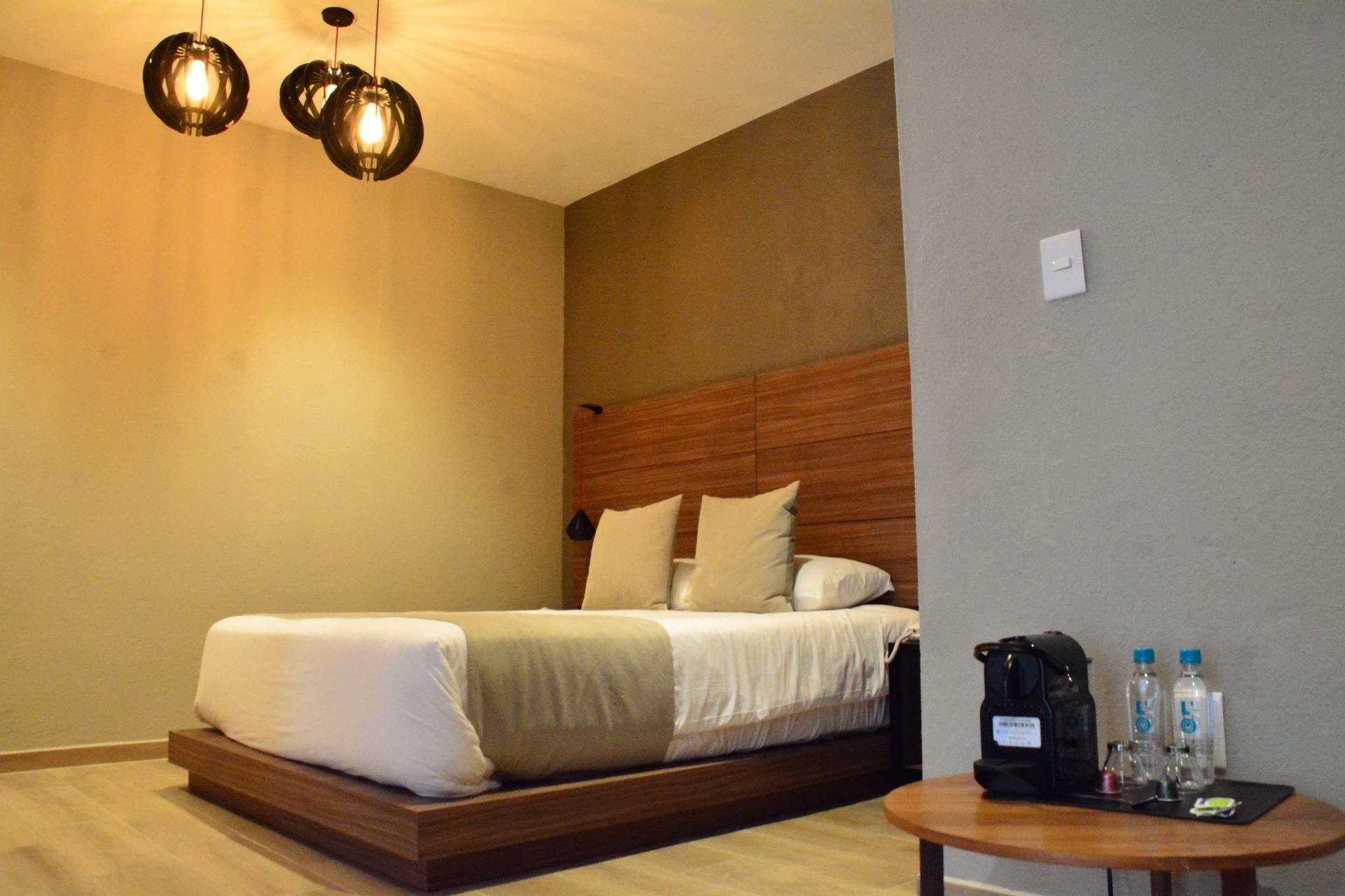 Dormitorio con cama, cabecero de madera, lámparas redondas y mesa con cafetera.