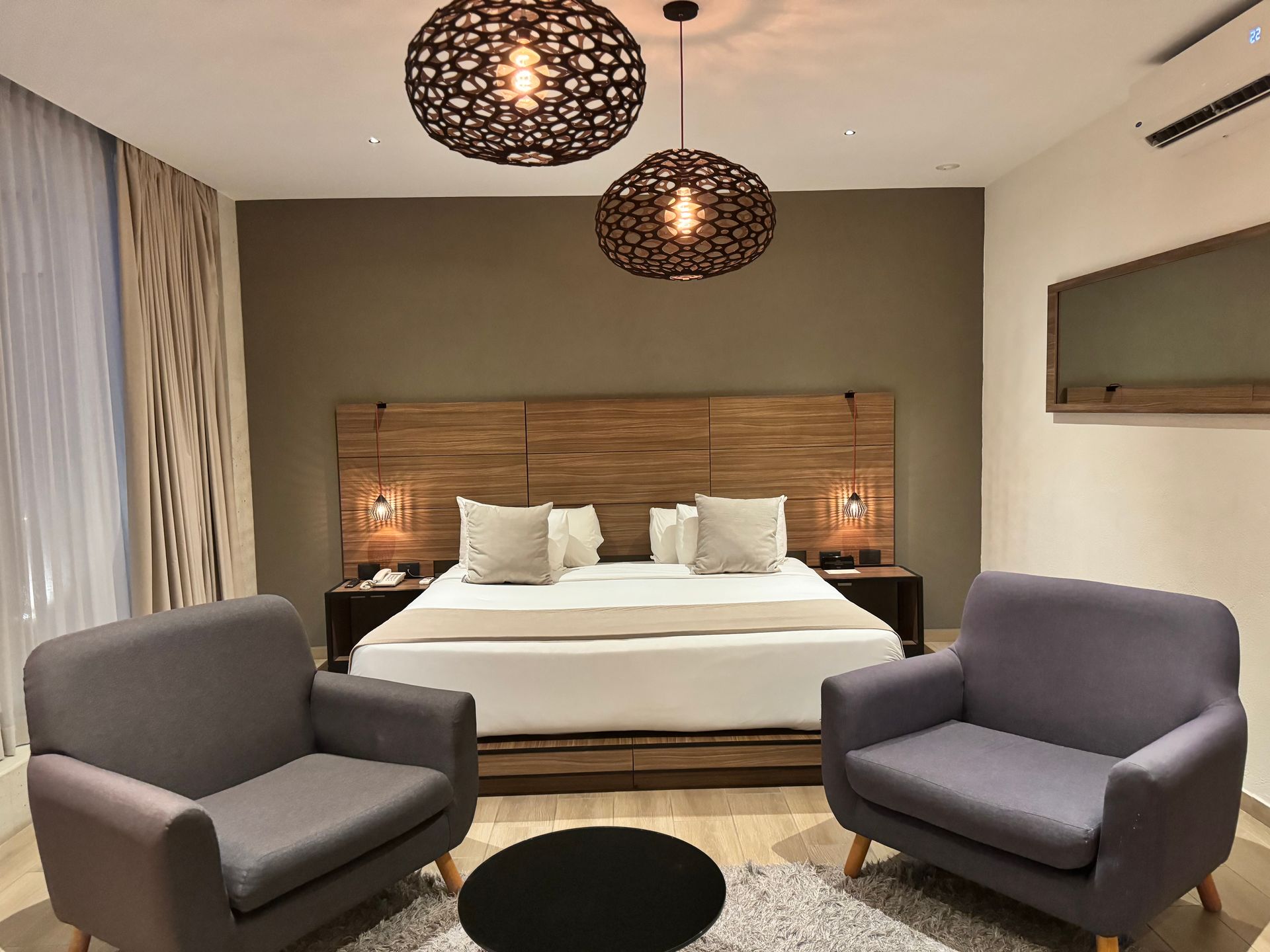 Dormitorio con cama, dos sillones grises, mesa redonda negra, luces decorativas, pared beige.