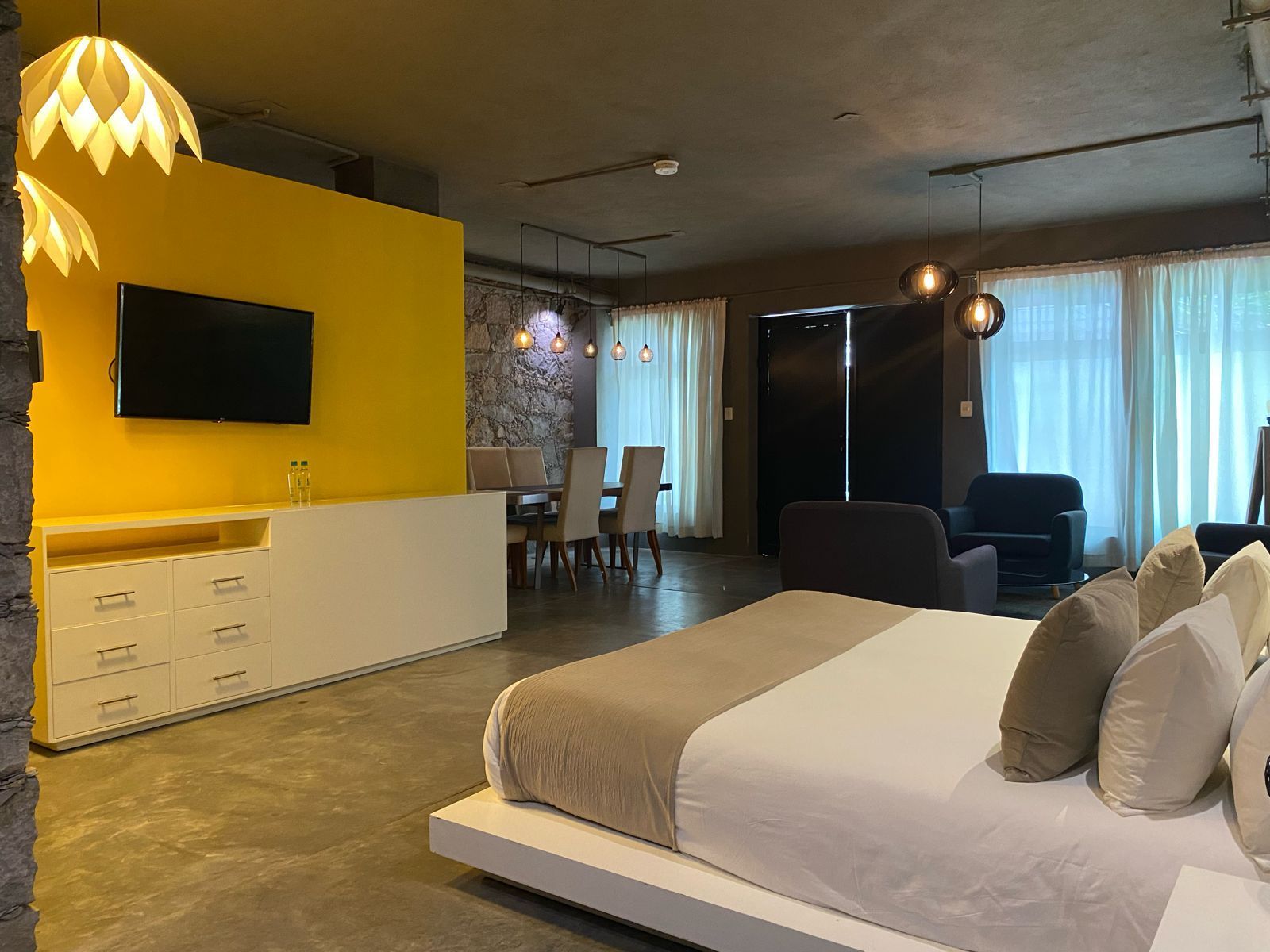 Suite moderna con pared con acento amarillo, TV, cama, mesa de comedor y zona de estar.