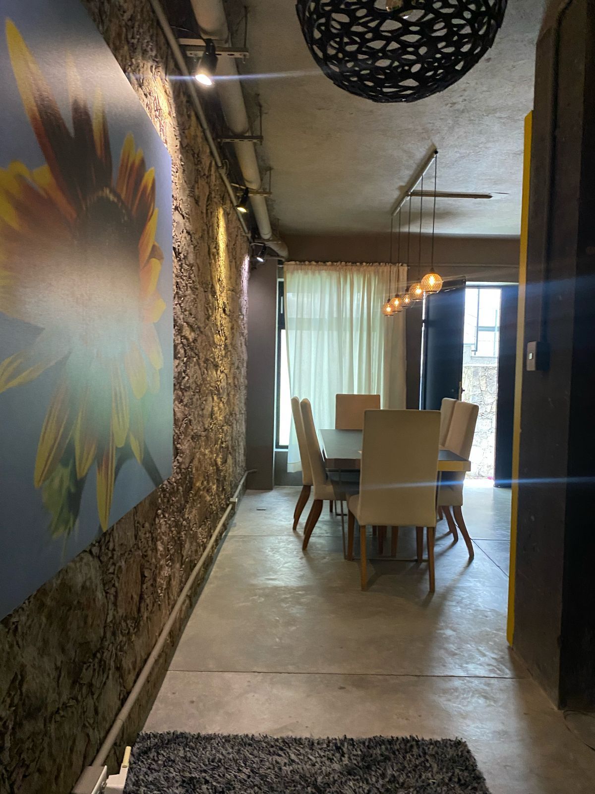 Pasillo con pared de piedra, cuadro de girasol, mesa de comedor con sillas y puerta oscura abierta.