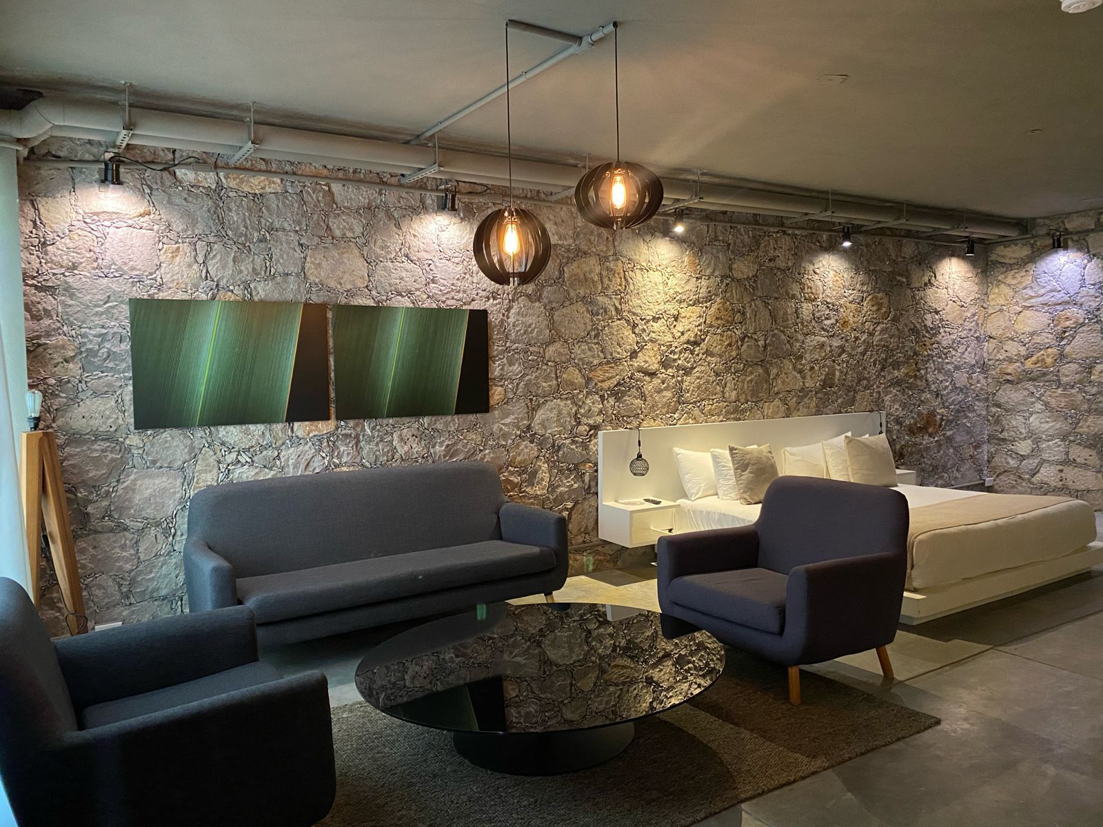 Habitación con paredes de piedra, muebles grises, una cama y obras de arte. Dos lámparas colgantes de globo.