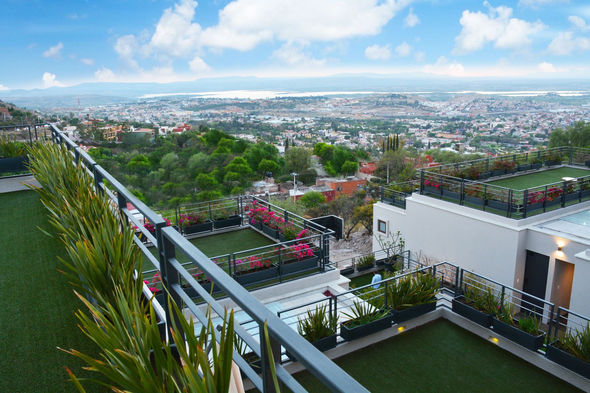 Vista de una ciudad desde una terraza en la azotea con césped verde y plantas con flores; cielo azul.