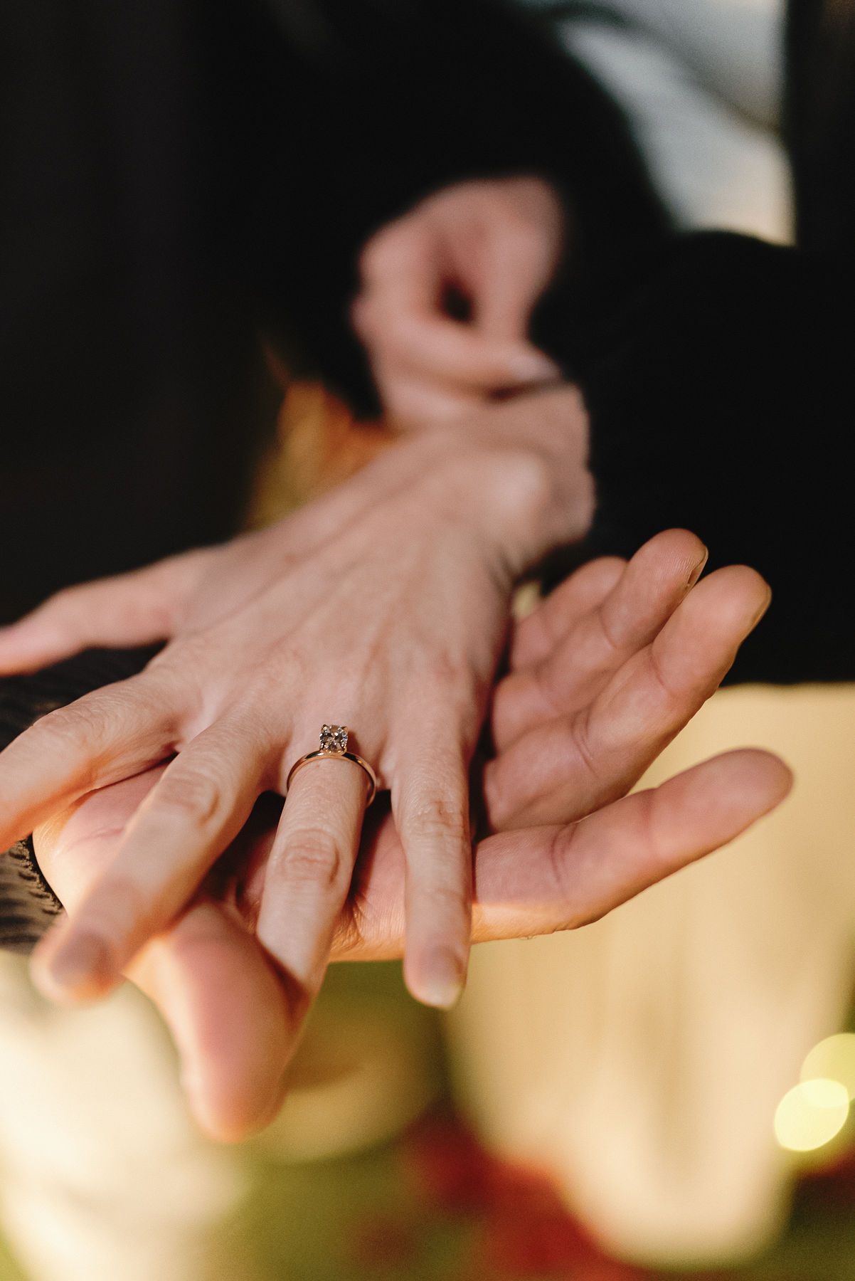 La mano de una persona con un anillo de compromiso encima de la mano de otra persona, contra un fondo borroso.