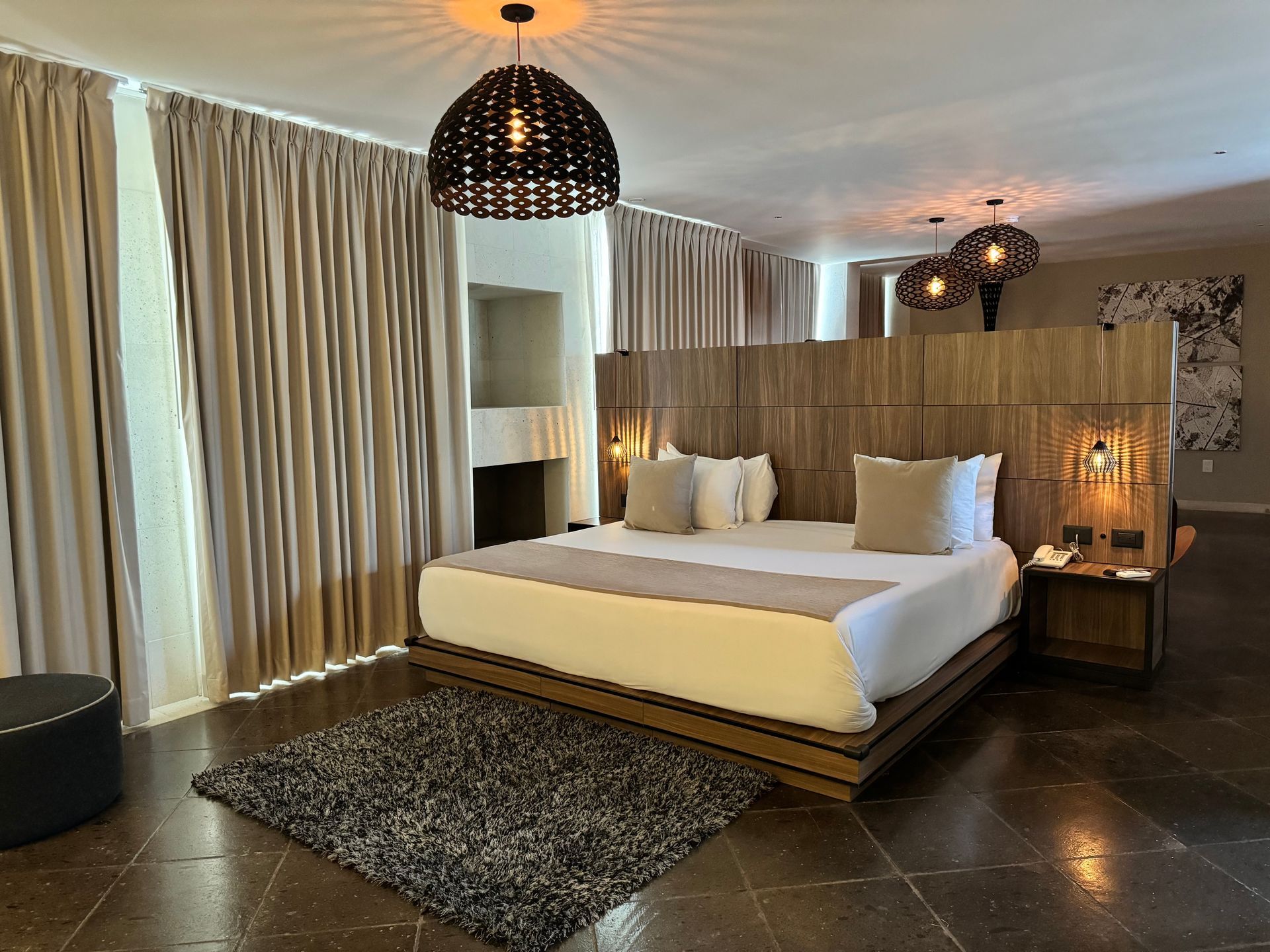 Dormitorio con cama grande, tonos neutros, detalles en madera, iluminación decorativa y alfombra.