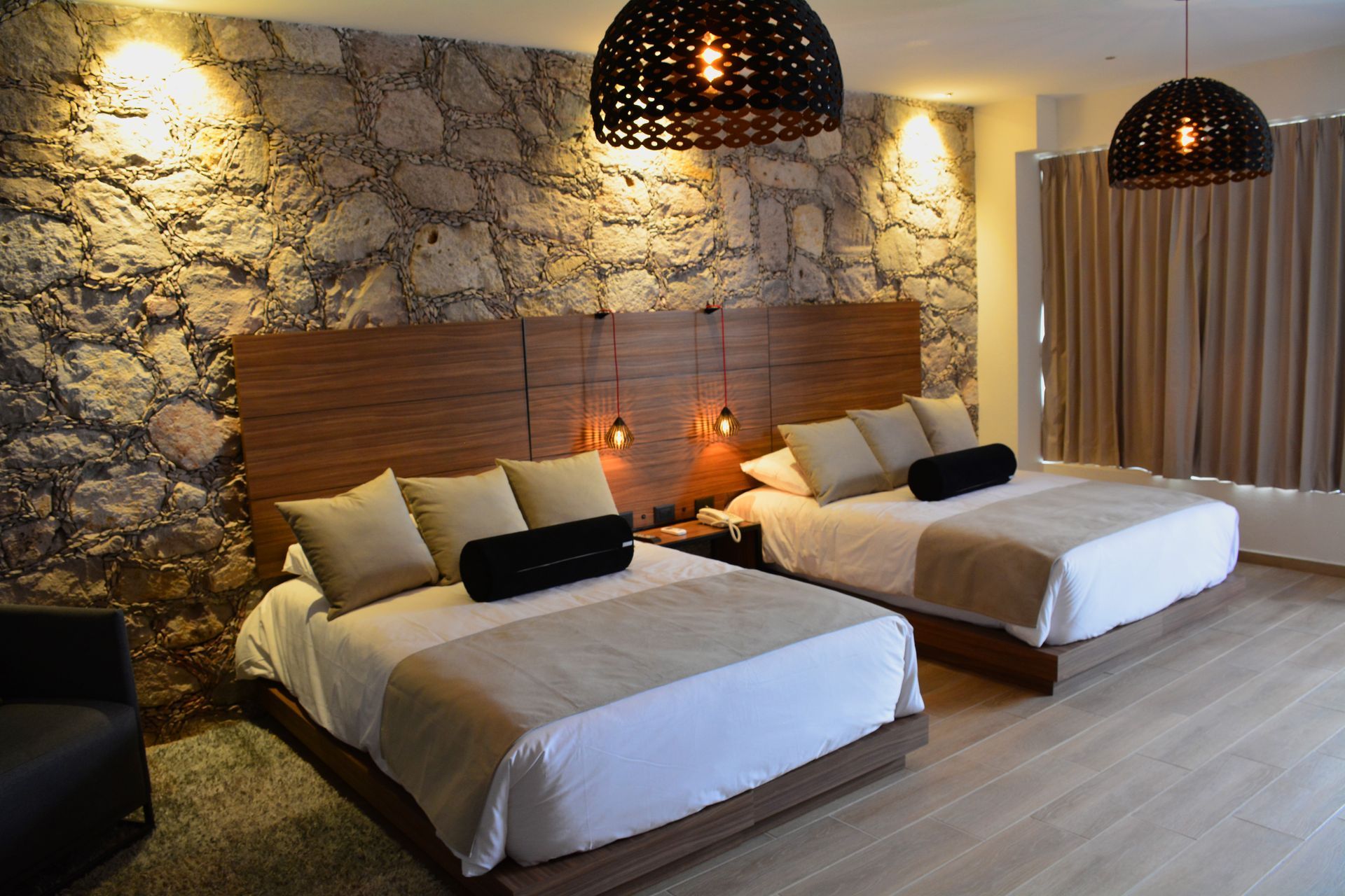 Habitación de hotel con dos camas queen, pared de piedra, cabecero de madera, lámparas colgantes.