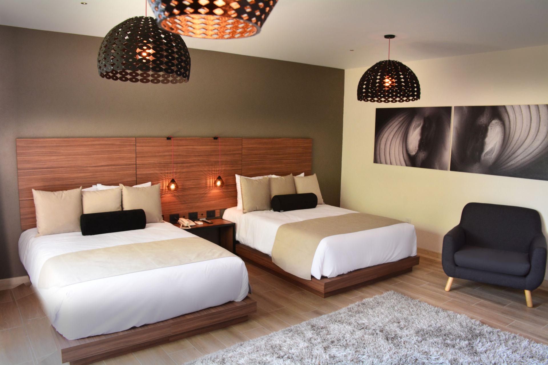 Habitación de hotel con dos camas, cabecero de madera, iluminación moderna y sillón gris.