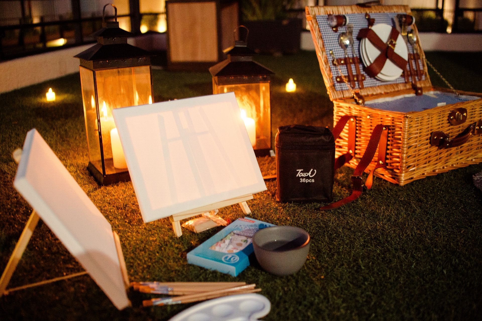 Instalación de pintura al aire libre con linternas, canasta de picnic, lienzos en blanco y materiales de arte.