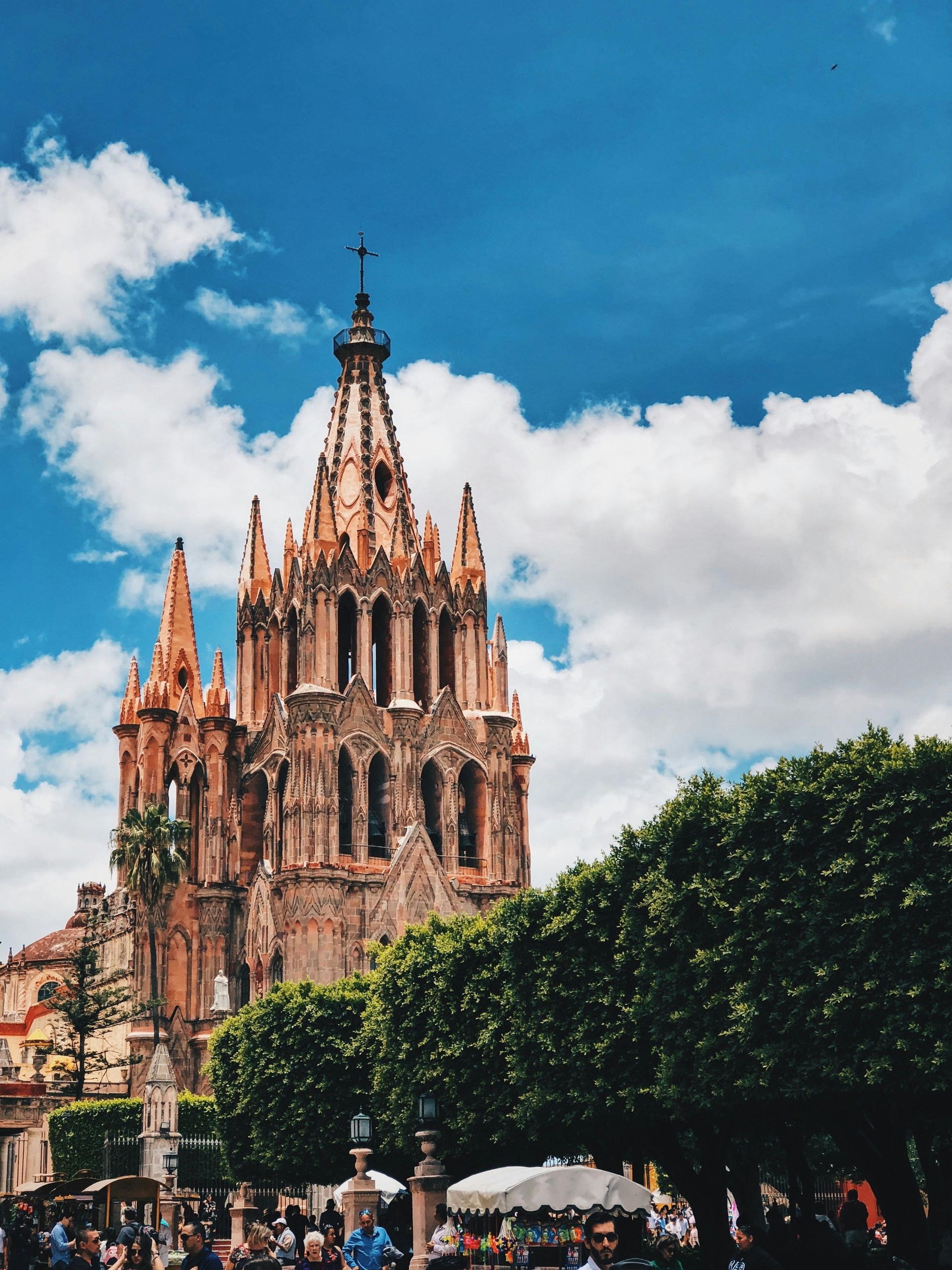 Iglesia gótica contra un cielo azul con nubes esponjosas. Árboles en primer plano, gente visible abajo.
