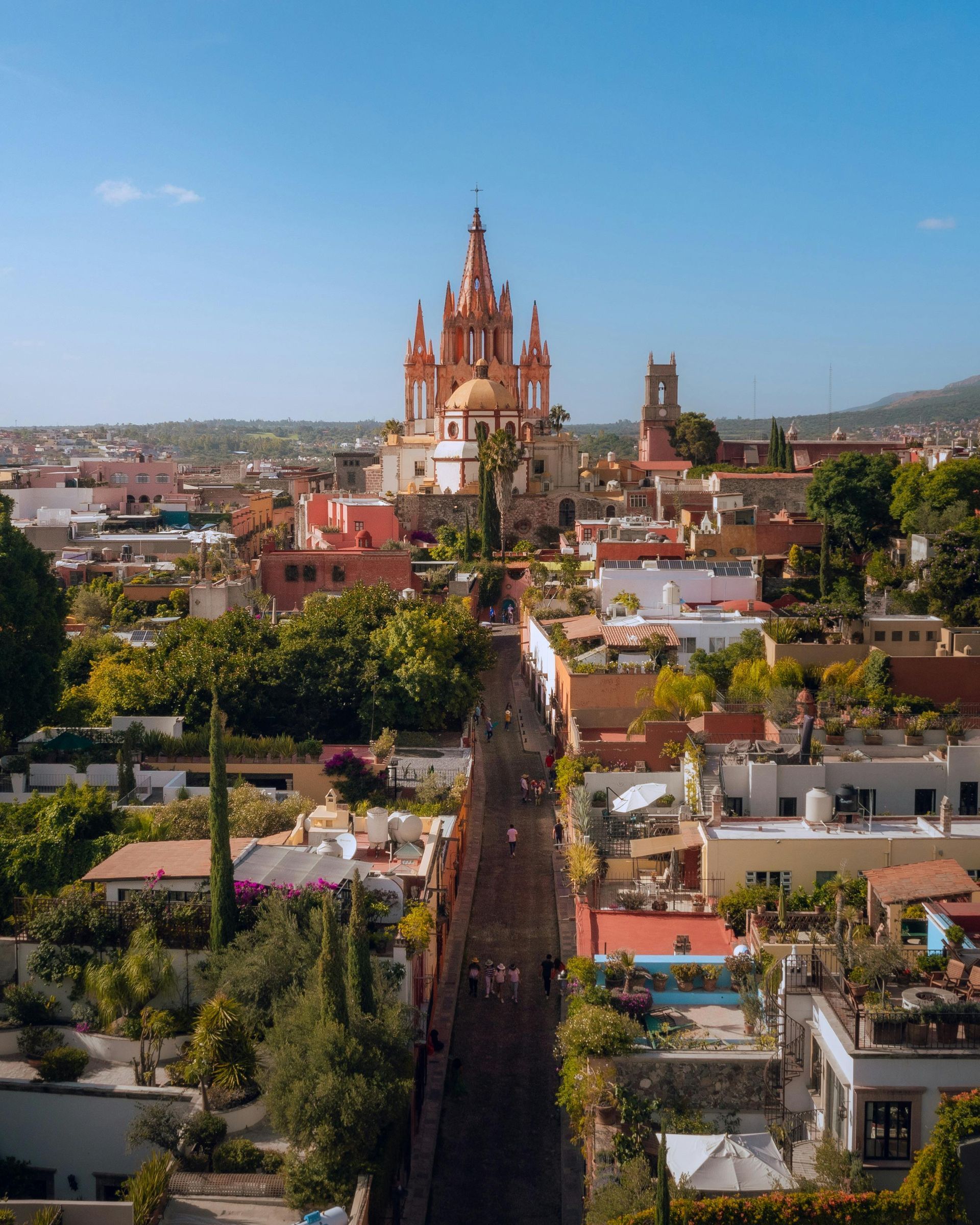 Vista de una calle de la ciudad que conduce a una iglesia rosa con torres, edificios y árboles bajo un cielo azul.