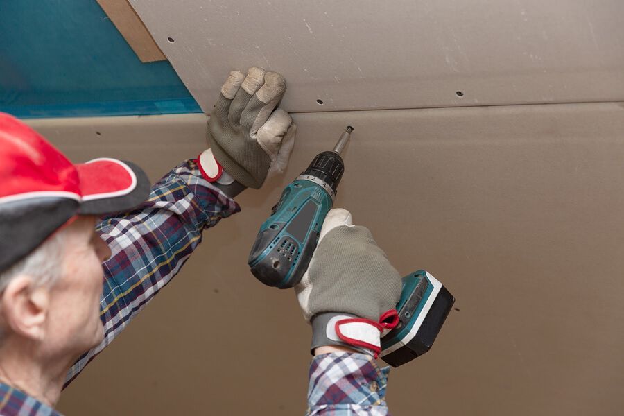 Person using a drill to attach drywall to a ceiling. They wear work gloves and a plaid shirt.