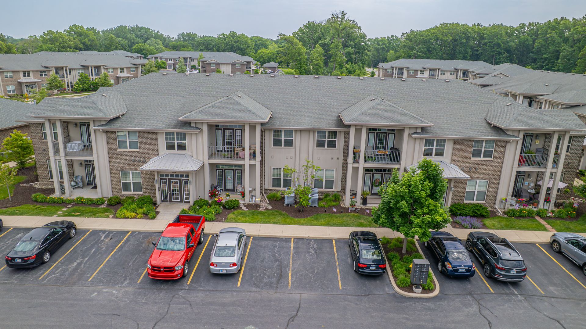 Apartment building with cars parked in front. Gray roof, brick facade, trees in the background.
