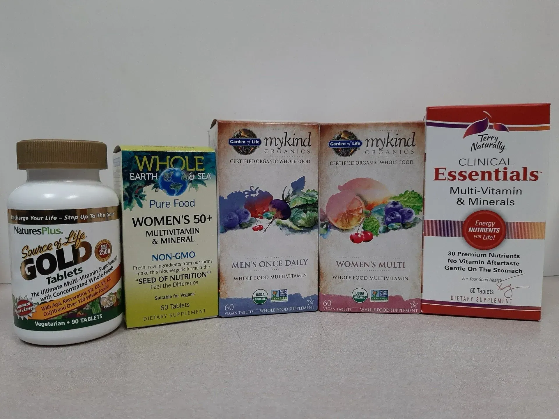 A line of five multivitamin supplement containers, including Garden of Life brand bottles and boxes, on a flat surface.