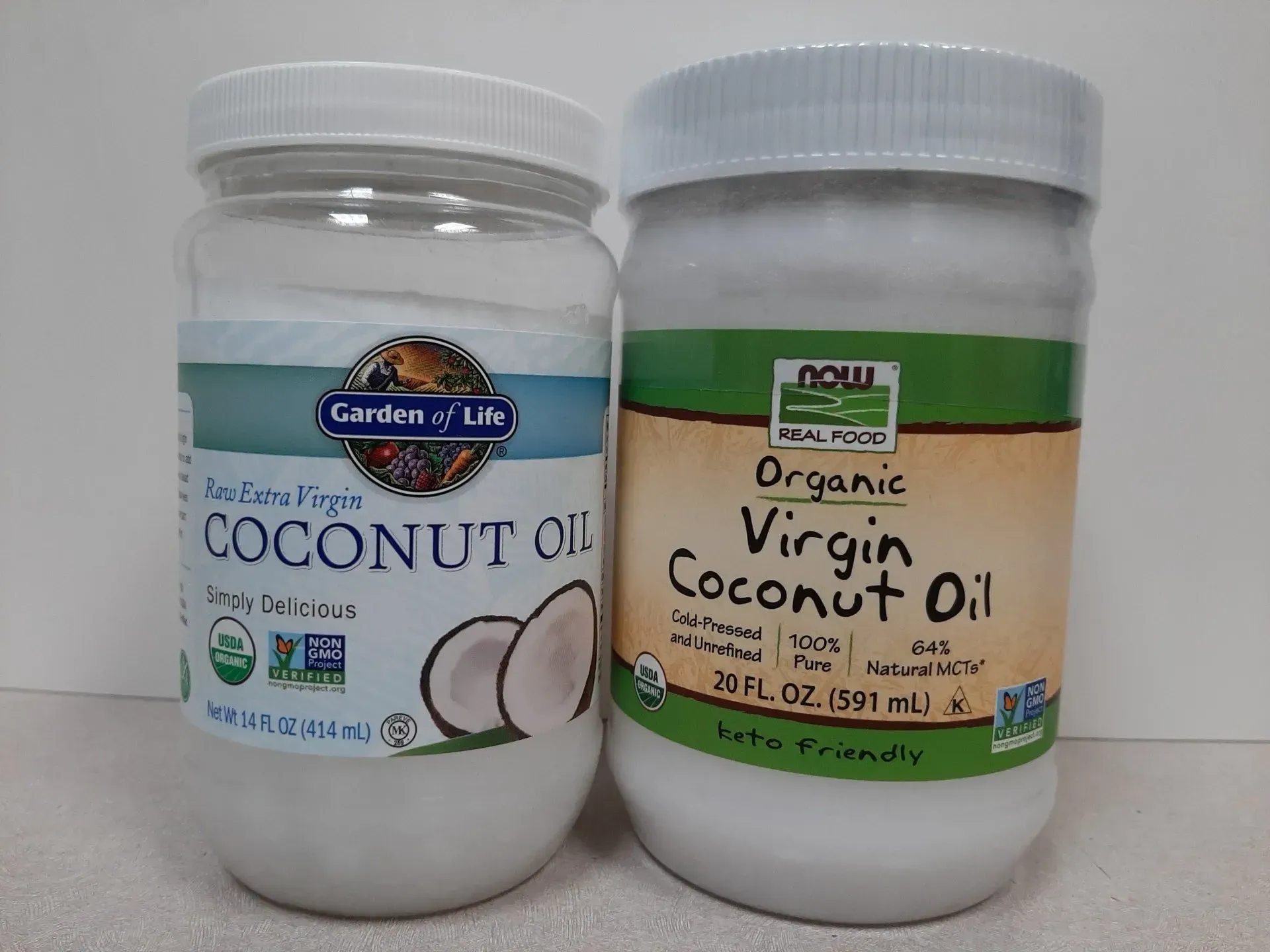 Two jars of organic virgin coconut oil from the brands Garden of Life and NOW Real Food sit side-by-side on a white surface.