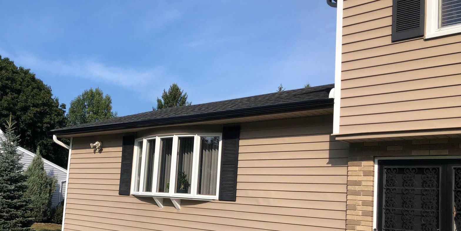 Black gutters installed on a tan house with a black roof. The new gutter is installed above a pretty bay window.