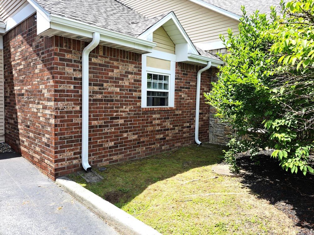 Freshly installed gutters on a brick house. Two gutter spouts come down the house on either side of a small window.