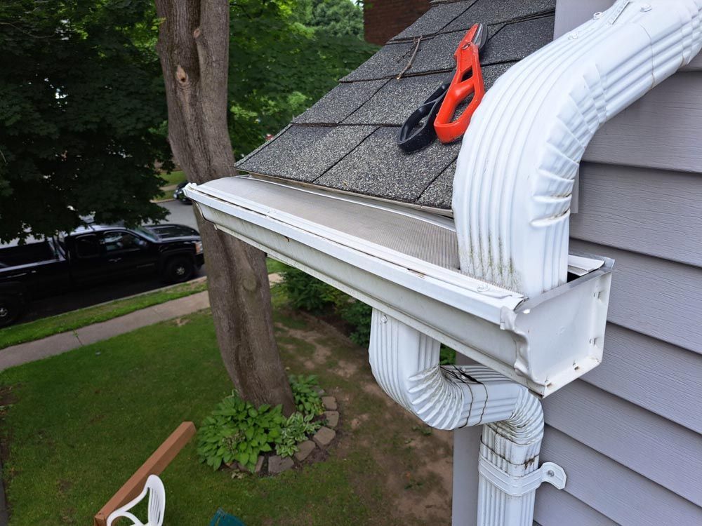 A white -colored gutter system in process of installation on a bluish gray house. A tool is partially visible on the roof.