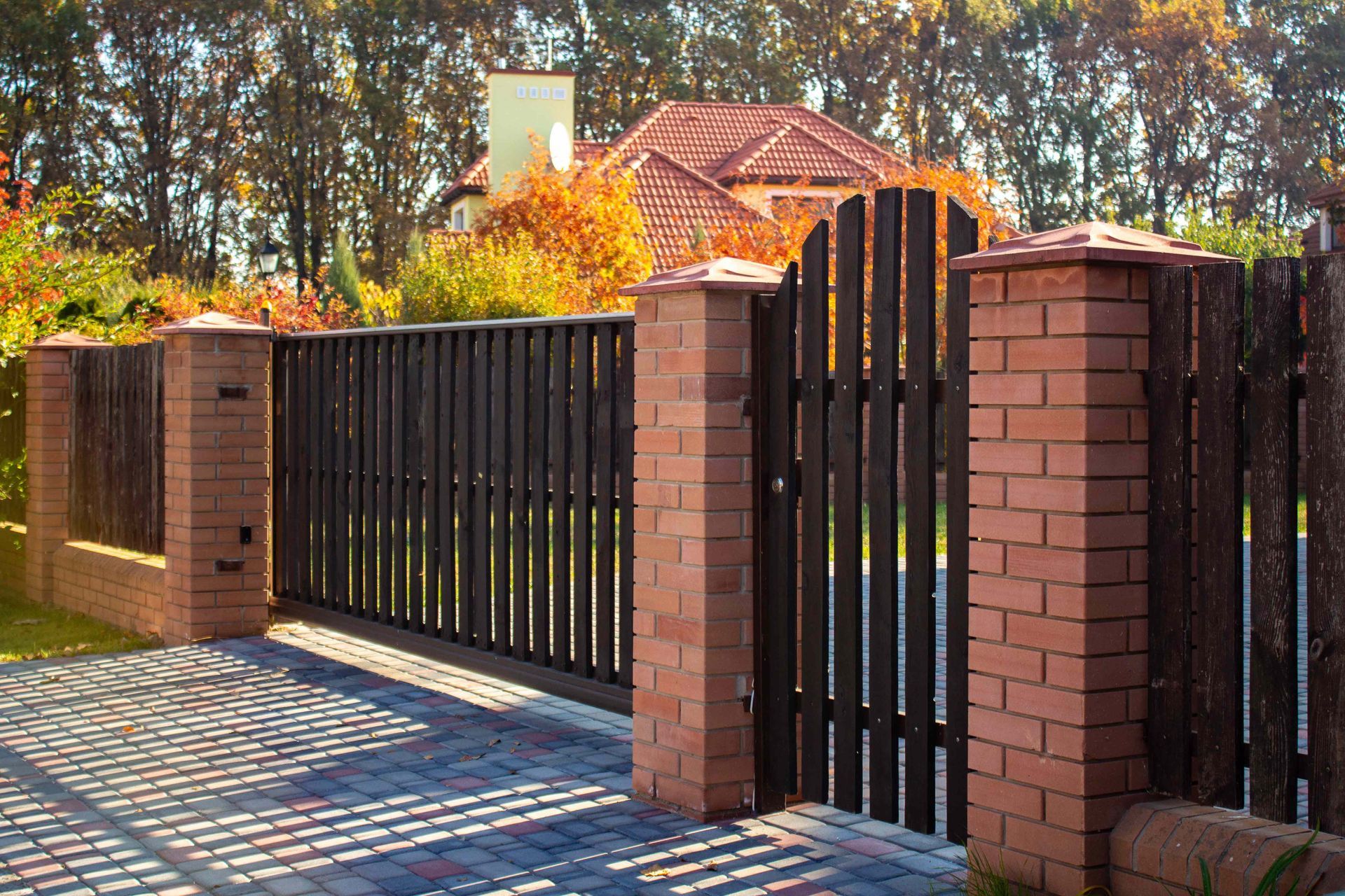 Brick-and-black metal driveway gate beside a brick fence, with autumn trees in the background