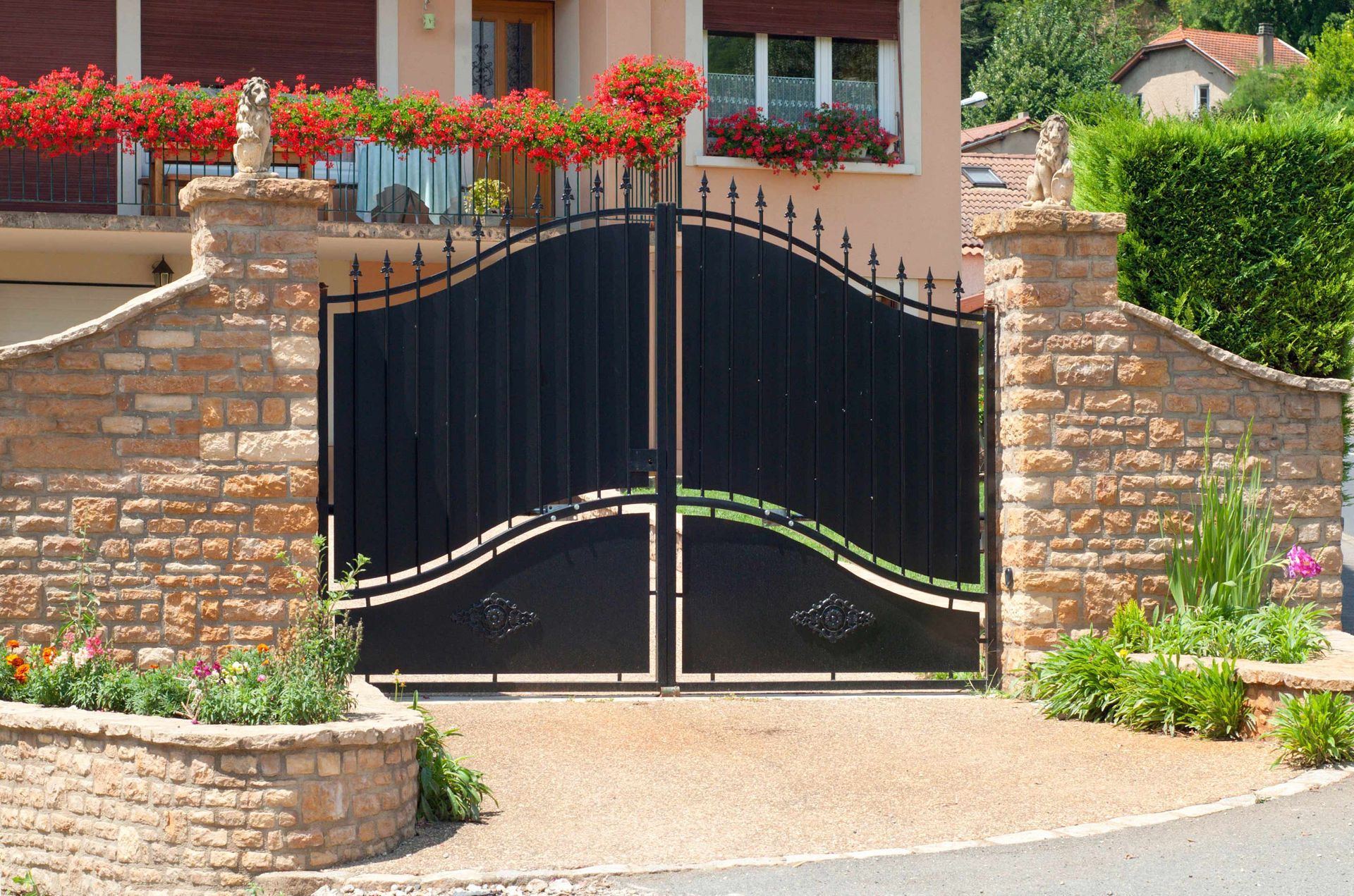 Black wrought-iron gate in a stone wall, with flowers and a house in the background. Black wrought-iron gate in a stone wall, with flowers and a house in the background.