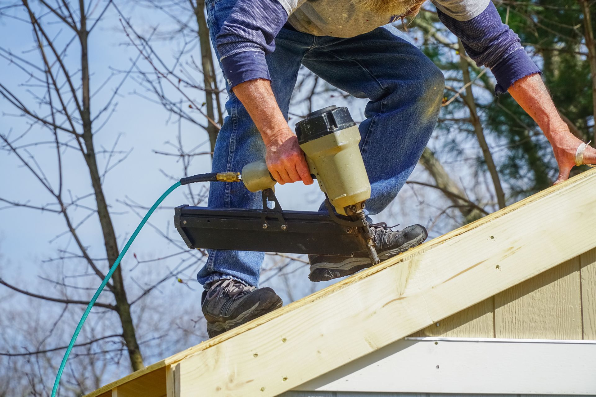 Worker using a nail gun on a roof, fastening light-colored boards outdoors. Worker using a nail gun on a roof, fastening light-colored boards outdoors.